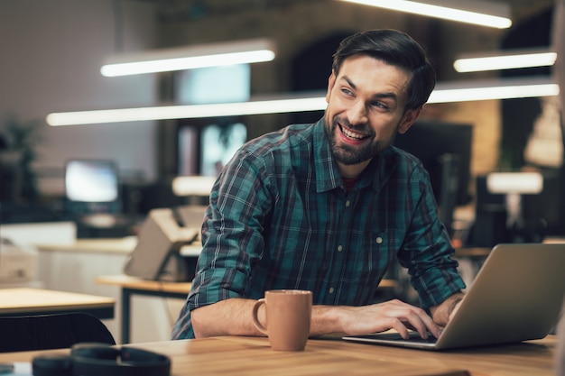 Fröhlicher gutaussehender Mann, der in Freizeitkleidung am Laptop arbeitet und mit einem Lächeln wegschaut