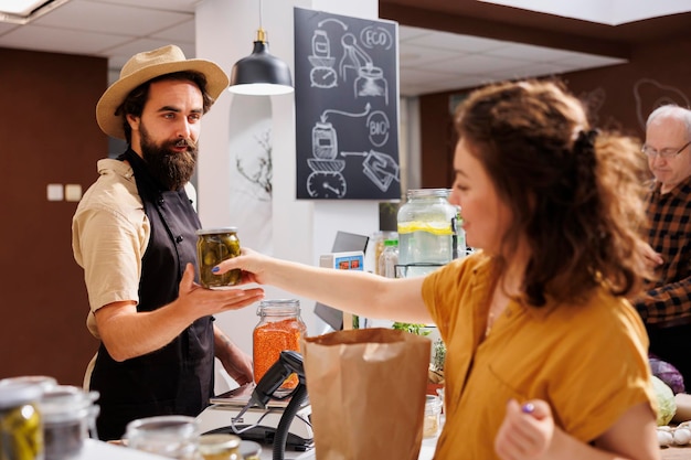 Fröhlicher Händler an der Kasse in einem Zero-Waste-Shop, der vegane Produkte an umweltbewusste Kunden verkauft. Frau kauft gesunde Lebensmittel in einem örtlichen Laden mit geringem CO2-Fußabdruck