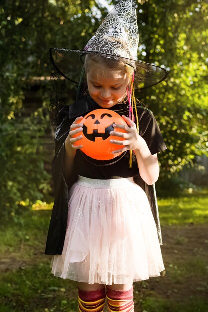 Fröhlicher Halloween-Tag Kleines Mädchen in einem Hexenkostüm hält einen Eimer mit Süßigkeiten und schaut auf Süßigkeiten