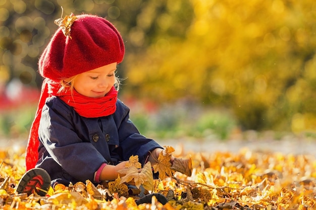 Fröhlicher Herbst Ein kleines Mädchen in einer roten Baskenmütze spielt mit fallenden Blättern und lacht