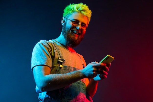 Fröhlicher Hipster-Mann mit Bart und grünen Haaren, der mit zahnigem Lächeln in die Kamera schaut und das Handy in den Händen hält, gut gelaunt ist, hat tolle Neuigkeiten. Buntes Neonlicht, Studioaufnahme im Innenbereich.