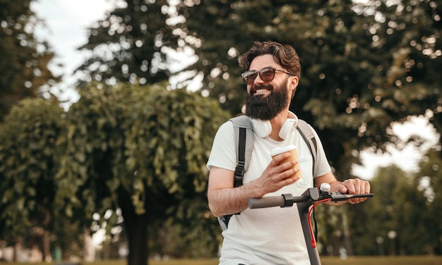 Fröhlicher Hipster mit Heißgetränk auf Roller