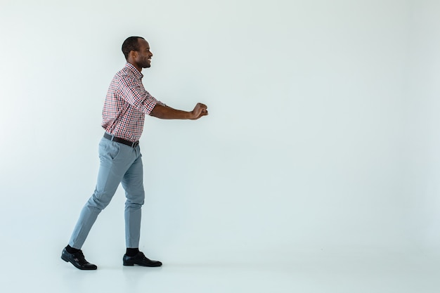 Fröhlicher hübscher afroamerikanischer Mann, der einen Einkaufswagen gegen weiße Wand hält