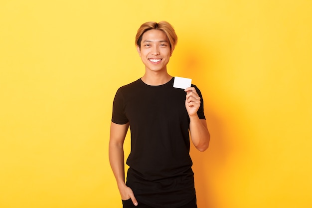 Fröhlicher hübscher asiatischer Kerl, der erfreut lächelt und Kreditkarte zeigt