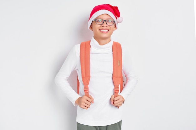 Fröhlicher hübscher junger asiatischer Student in einer Weihnachtsmütze, der ein T-Shirt mit einem Rucksack trägt und selbstbewusst in die Kamera schaut, isoliert auf weißem Hintergrund