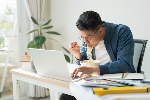 Fröhlicher Ingenieur mit Dessert, der vor dem Computermonitor sitzt und Nudeln isst