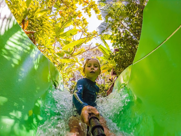 Fröhlicher Junge auf Wasserrutsche in einem Schwimmbad, der sich während der Sommerferien in einem schönen