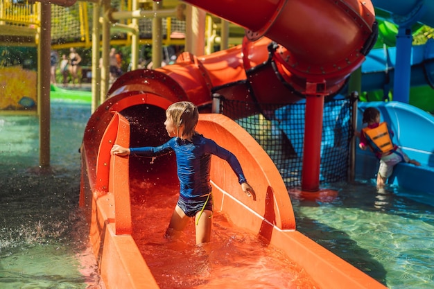 Fröhlicher Junge auf Wasserrutsche in einem Schwimmbad, der sich während der Sommerferien in einem schönen