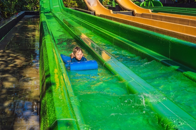 Fröhlicher Junge auf Wasserrutsche in einem Schwimmbad, der sich während der Sommerferien in einem schönen