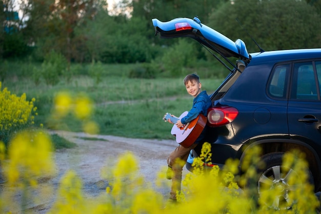 Fröhlicher Junge, der Gitarre spielt und die Sommerzeit genießt, während er auf dem offenen Kofferraum des Autos sitzt. Reise-, Urlaubs- und Abenteuerkonzept.
