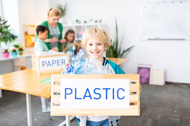 Fröhlicher junge. fröhlicher blonder süßer junge, der nach dem ökologieunterricht eine kiste mit plastik hält