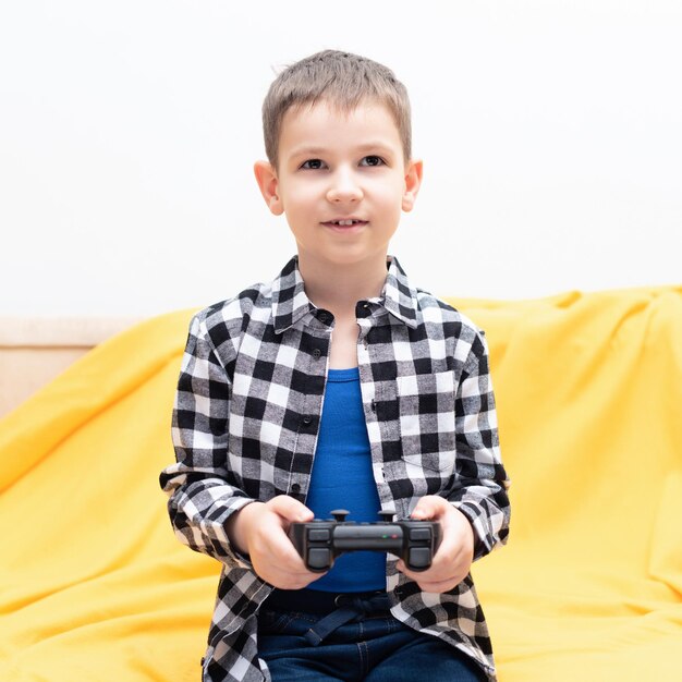 Fröhlicher Junge im karierten Hemd, der auf der Couch sitzt und mit einem schwarzen Joystick in den Händen das Videospiel spielt Videospiele zu Hause spielen Konzept