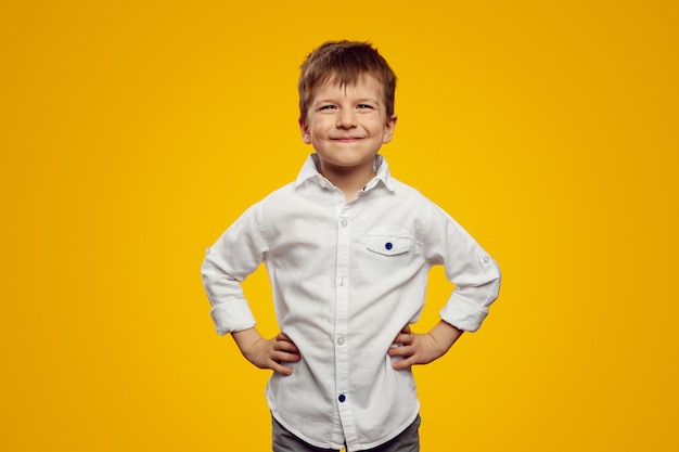 Fröhlicher Junge im weißen Hemd lächelt, während er die Hände auf der Taille hält, vor gelbem, hellem Hintergrund