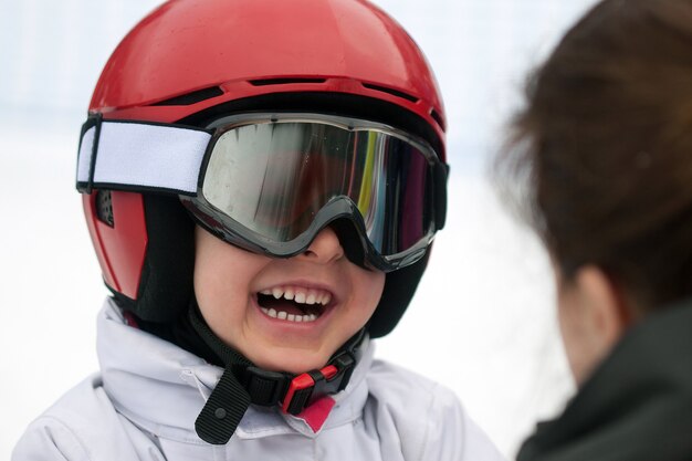 Fröhlicher Junge in rotem Helm, Skibrille und weißer Jacke, der zu seiner Mutter lächelt. Wintersport, junger Skifahrer, schneebedeckter Hintergrund