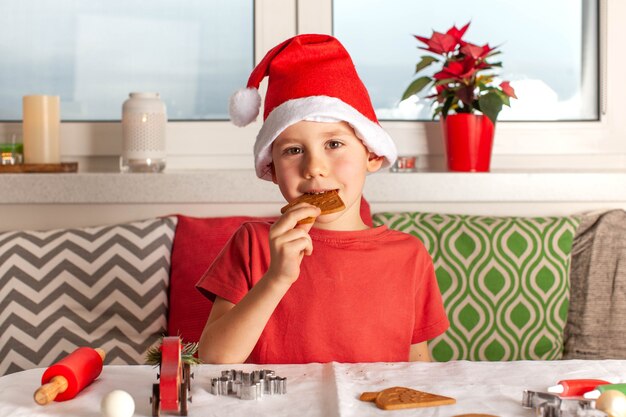 Fröhlicher Junge in Weihnachtsmann-Hut isst Weihnachtslebkuchen, der Neujahrslebkuchen kocht