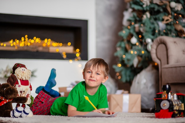 Fröhlicher Junge ist ein Brief an den Weihnachtsmann, in der Nähe des Weihnachtsbaumes. Glückliche Kindheit, Zeit für die Erfüllung von Wünschen.