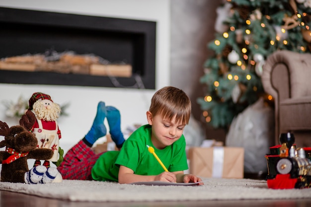 Fröhlicher Junge ist ein Brief an den Weihnachtsmann, in der Nähe des Weihnachtsbaumes. Glückliche Kindheit, Zeit für die Erfüllung von Wünschen.