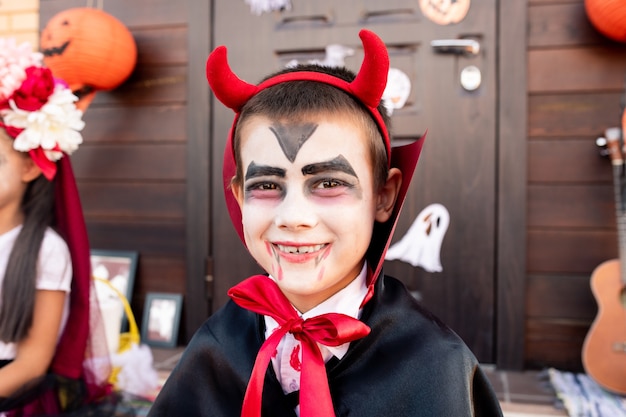 Fröhlicher Junge mit gemaltem Gesicht im Kostüm des Teufels, der vor Kamera gegen Freund sitzt, der Spaß hat