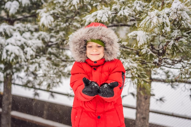 Fröhlicher Junge spielt mit Schnee Nettes Kind, das Schnee in einem Winterpark wirft Fröhliche Winterferien Wintermode