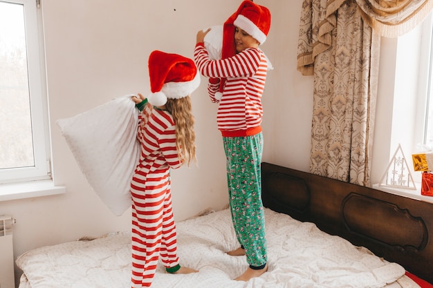 Fröhlicher Junge und Mädchen in Weihnachtsmützen am Weihnachtsmorgen spielen und kämpfen mit Kissen im Bett