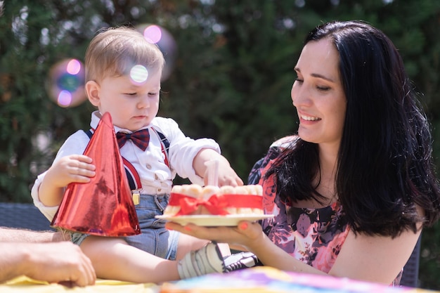 Fröhlicher Junge und Mutter auf seiner Geburtstagsfeier mit erster Torte und lustigem Hut und Seifenblasen