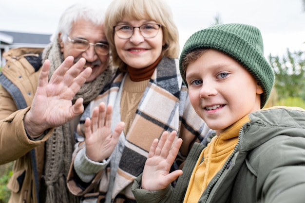 Fröhlicher Junge und seine liebevollen Großeltern winken mit den Händen und schauen lächelnd in die Smartphone-Kamera, während sie im Freien ein Selfie machen