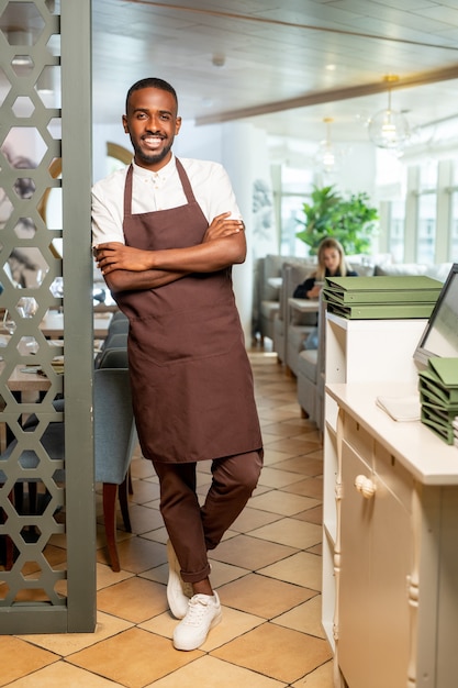 Fröhlicher junger afrikanischer Kellner in Arbeitskleidung, der sich an einem gedeckten Tisch im luxuriösen Restaurant an einen Sessel lehnt, während er auf neue Gäste wartet?