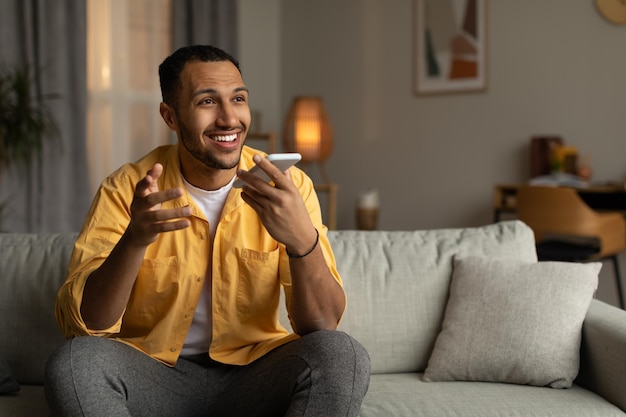 Fröhlicher junger Afroamerikaner, der am Handy spricht und zu Hause auf dem leeren Raum auf dem Sofa sitzt