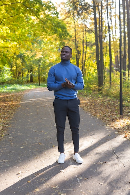 Fröhlicher junger Afroamerikaner junger Mann in den stilvollen Kleidern geht im Herbstpark am sonnigen warmen Herbsttag