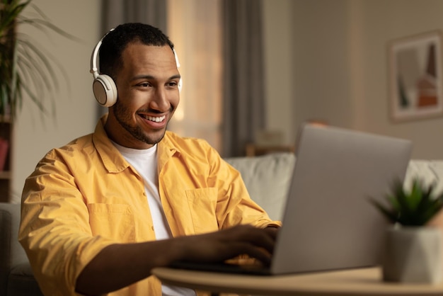 Fröhlicher junger afroamerikanischer Mann mit Laptop, der zu Hause einen Remote-Job mit Kopfhörern hat