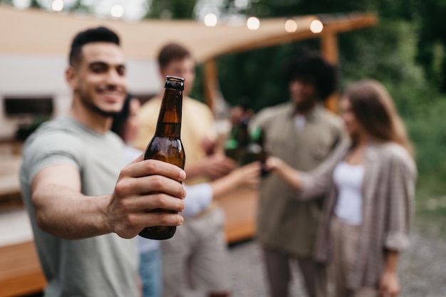 Fröhlicher junger arabischer Kerl mit diversen Freunden, die mit Bierflaschen anstoßen, die Alkohol in der Nähe von Wohnmobil trinken