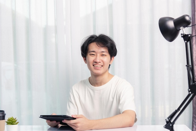 Fröhlicher junger asiatischer gutaussehender Mann arbeitet von zu Hause aus mit Brille, T-Shirt bequem, er lächelt und benutzt einen schwarzen, modernen, intelligenten digitalen Tablet-Computer, um E-Mails zu lesen oder auf dem Schreibtisch zu lernen