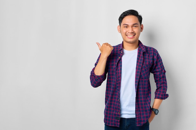 Fröhlicher junger asiatischer Mann im karierten Hemd, der mit dem Daumen auf das Werbebanner des Kopierraums zeigt, das auf weißem Hintergrund isoliert ist