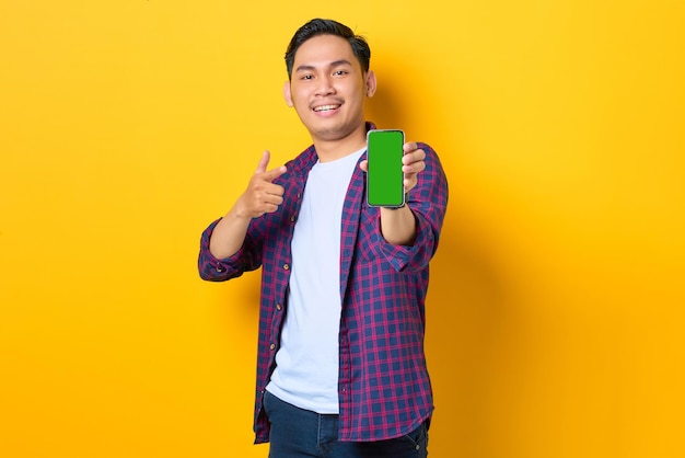 Fröhlicher junger asiatischer Mann im karierten Hemd mit Smartphone mit leerem Bildschirm isoliert auf gelbem Hintergrund