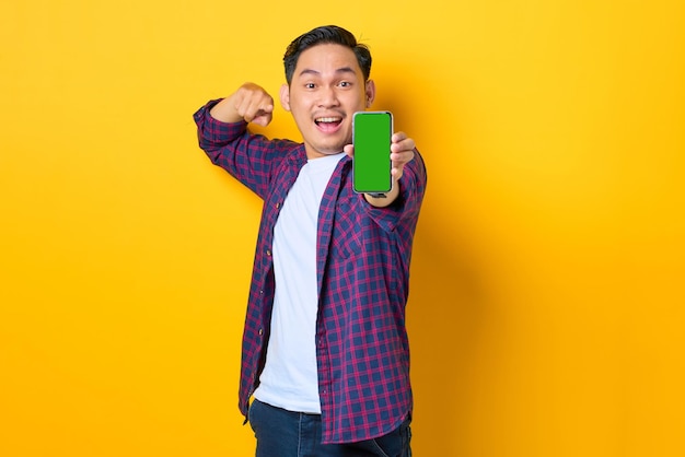 Fröhlicher junger asiatischer Mann im karierten Hemd mit Smartphone mit leerem Bildschirm isoliert auf gelbem Hintergrund