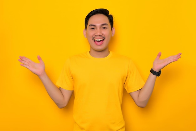 Fröhlicher junger asiatischer Mann in lässigem T-Shirt, der die Hände ausbreitet und Spaß daran hat, die Kamera isoliert auf gelbem Hintergrund zu betrachten Lifestyle-Konzept für Menschen