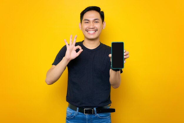 Fröhlicher junger asiatischer Mann in legerem T-Shirt mit Handy und leerem Bildschirm mit ok-Geste isoliert auf gelbem Hintergrund Lifestyle-Konzept für Menschen