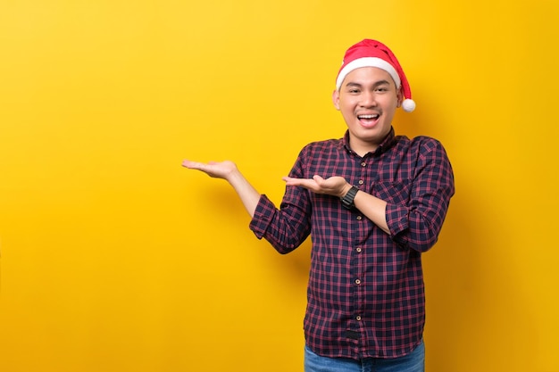 Fröhlicher junger asiatischer Mann mit Weihnachtsmütze, der leeren Raum auf der Handfläche über gelbem Studiohintergrund zeigt Frohes neues Jahr 2023 Feier frohes Feiertagskonzept