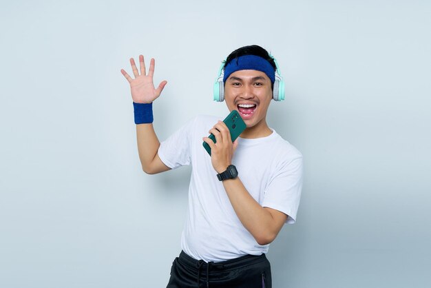 Fröhlicher junger asiatischer Mann, sportlicher Fitness-Trainer in blauem Stirnband und weißem T-Shirt mit Kopfhörern, die Musik hören, singen Lied im Diktiergerät auf dem Mobiltelefon vor weißem Hintergrund