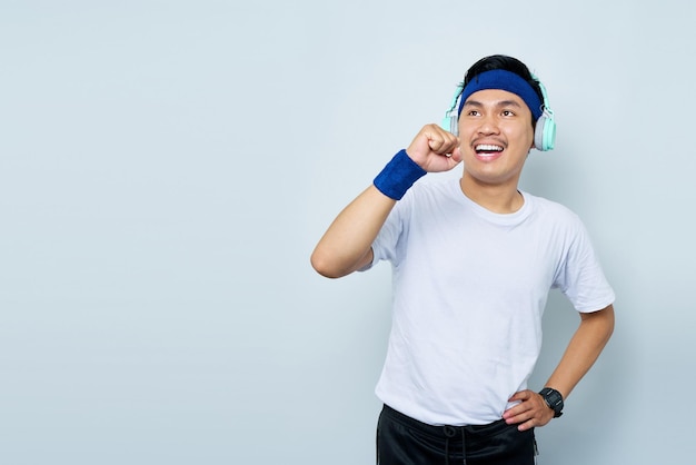 Fröhlicher junger asiatischer Mann, sportlicher Fitness-Trainer in blauem Stirnband und weißem T-Shirt mit Kopfhörern, hört Musik, singt Lied im Mikrofon, isoliert über weißem Hintergrund