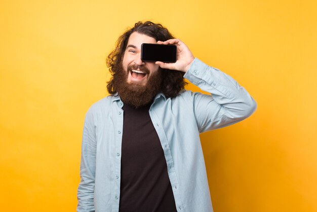 Fröhlicher junger bärtiger mann mit langen haaren, der smartphone über dem auge hält und über gelbem hintergrund lächelt