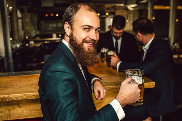 Fröhlicher junger bärtiger Mann schauen auf Kamera und lächeln. Er hält einen Krug Bier in der Hand. Zwei andere Leute stehen dahinter und reden.