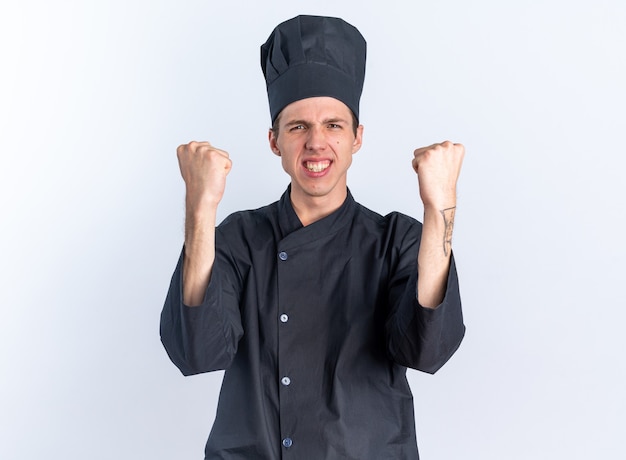 Fröhlicher junger blonder männlicher Koch in Kochuniform und Mütze, der die Kamera anschaut und die Ja-Geste isoliert auf weißer Wand macht