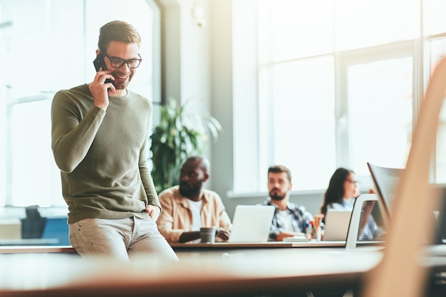 Fröhlicher junger Büroangestellter, der am Arbeitsplatz telefoniert