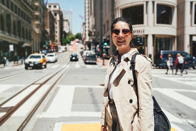 Fröhlicher junger College-Student mit Sonnenbrille und lächelnder Gesichtskamera, der an einem sonnigen Tag auf dem Zebrastreifen spazieren geht. Die Leute fahren mit ihren Privatautos zur Arbeit in San Francisco. Taxitransporte in der Stadt.