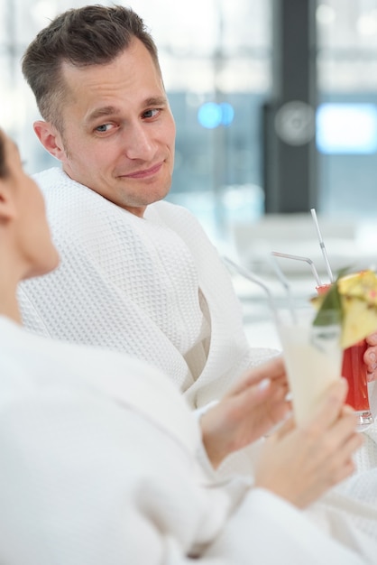 Fröhlicher junger, erholsamer Mann im weißen Bademantel, der seine Frau während des Gesprächs anschaut, während er auf Liegestühlen sitzt und Cocktails trinkt