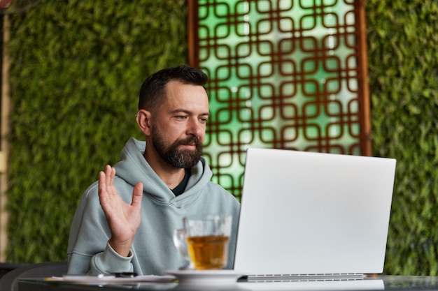 Fröhlicher junger erwachsener Mann in Freizeitkleidung im Café, der über Videoanrufe im Online-Geschäft spricht