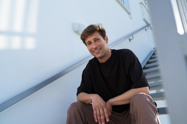 Fröhlicher junger erwachsener Mann in Sportkleidung sitzt auf der Treppe in der Nähe seines Hauses