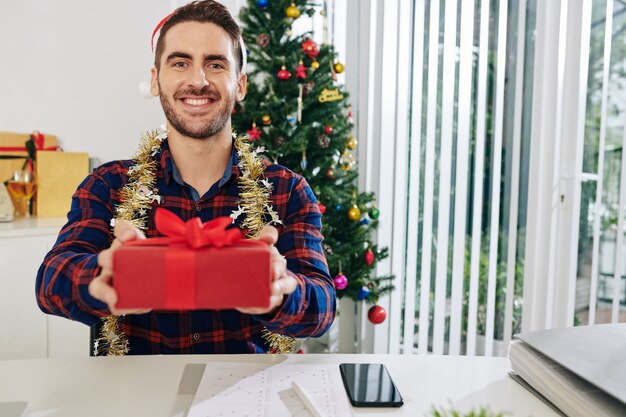 Fröhlicher junger Geschäftsmann, der ein verpacktes Weihnachtsgeschenk gibt, wenn er am Arbeitsplatz sitzt