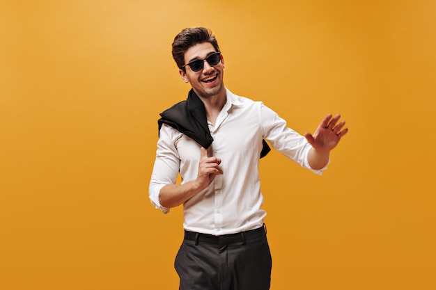 Fröhlicher junger gutaussehender Mann in Hosen, weißem Hemd, Sonnenbrille, der aufrichtig lächelt und eine schwarze Jacke an der orangefarbenen Wand hält.